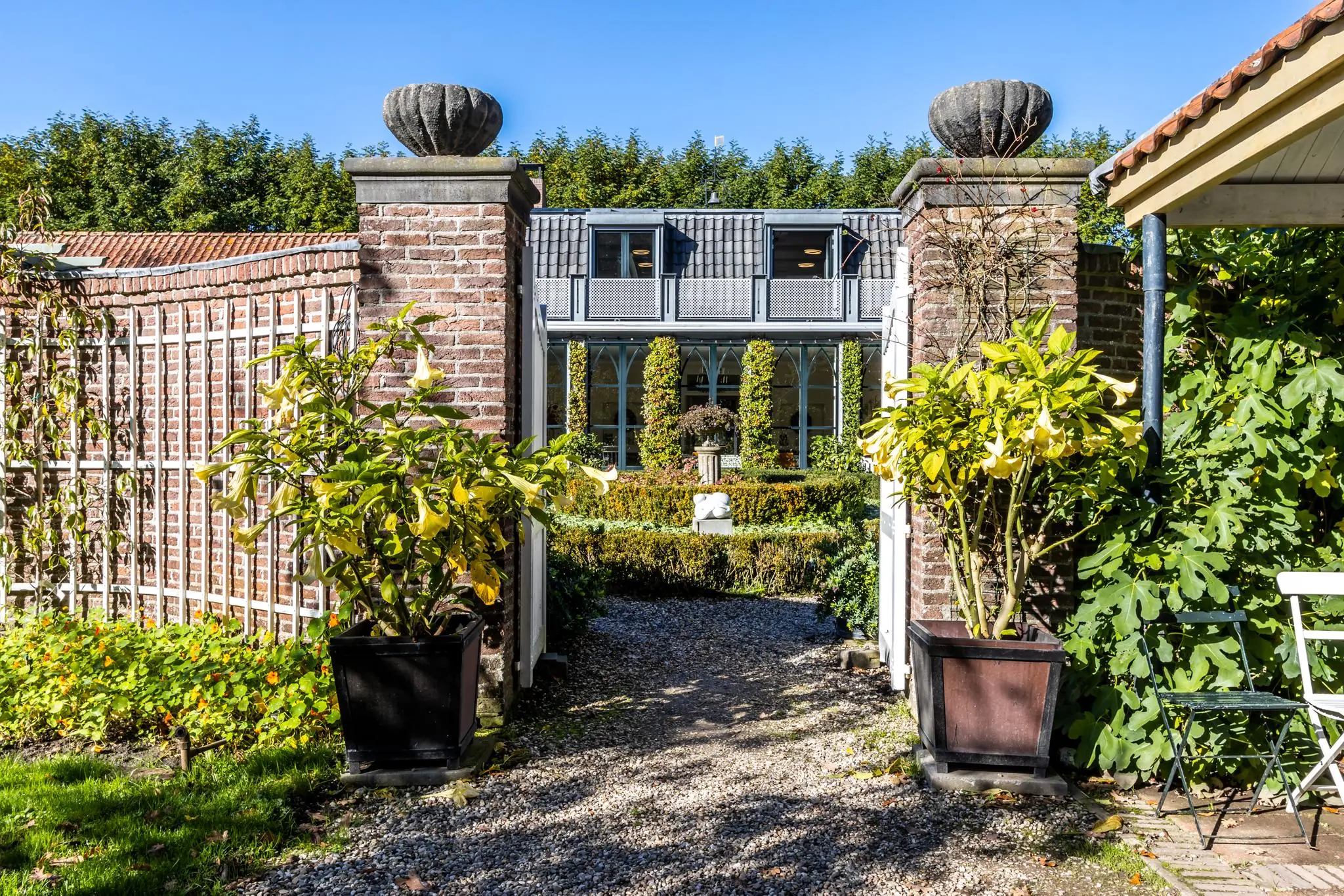 Toegangspoort met stenen pilaren en bloeiende planten leidt naar een fraai onderhouden tuin en een klassiek huis aan de Langbroekerdijk.